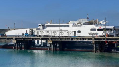 Ferry operator DFDS' Tarifa Jet boat docked in Jersey. The top half of the ferry is white and the lower half is dark blue. It is a sunny day.