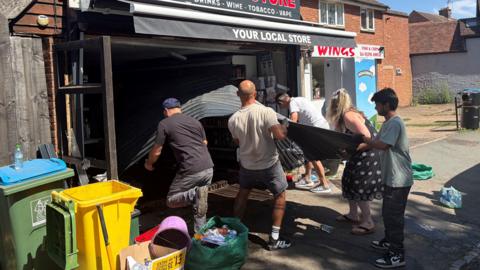 A group of people, helping to clear up a shop, holding onto a shutter. There are five people bending over items. There are bins to the left and bags full of items. The shop front looks dark.