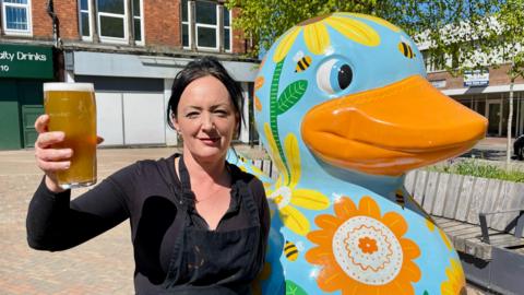 A woman with black hair, a black shirt and a black apron is holding a pint of lager aloft. She is stood beside a giant painted duck win a town square. The duck is blue, with an orange beak with flowers and bees painted on it. There are shops, benches, a tree and flower beds in the background.