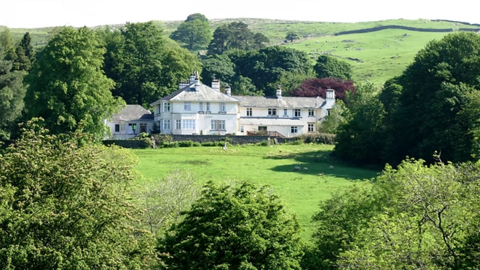 The large white building is two stories tall and has a grey roof with chimneys rising out of it. The building is nestled in a green field surrounded by trees. There are rolling hills in the background.