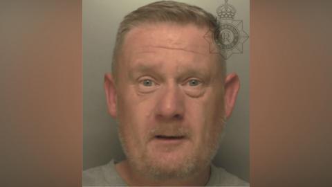 A custody image of Lawrence Worville who is wearing a grey sweatshirt as he stands against a grey background. He has light blue eyes, a beard and short light brown hair.