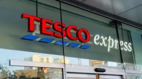 A Tesco Express sign, from a generic store, on glass above the entrance doors. "Tesco" is in red with blue dashes below it, and "express" is in white. 