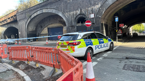 Police car parked across the road, barring it to traffic. Police tape is also across it. The corner also shows orange barriers around roadworks.