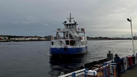 The Shields Ferry, a white and blue boat which has Pride of the Tyne written on the back of it. It has an indoor level, with an outdoor level above it. It is coming into one of the landings which is surrounded by a blue and white fence. One person is waiting by the barriers.