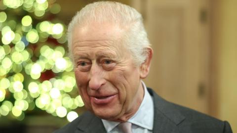 King Charles smiling at an event in the run-up to Christmas 2024, with a lit Christmas tree in the background. 