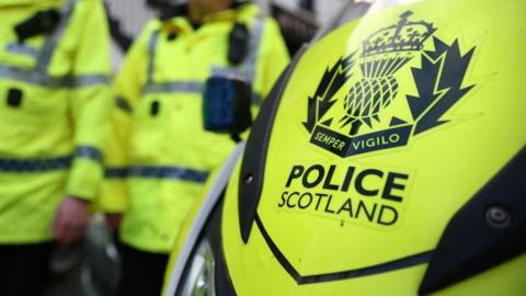 Police officers, in yellow jackets, in front of a Police Scotland sign on a vehicle.