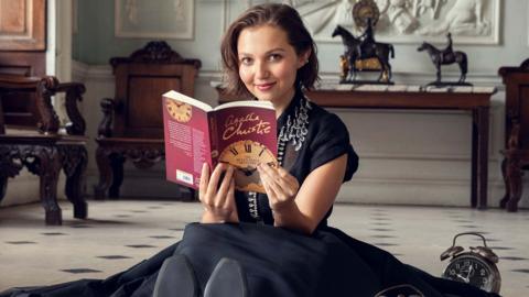 A young woman (Mia McKenna-Bruce) sitting on the floor as she reads an Agatha Christie novel. She's smiling up at the camera