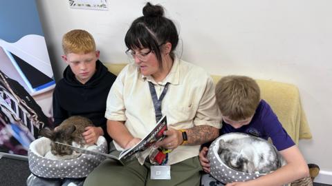 Two young boys and a woman sit on a sofa together reading a story. Both the boys have a rabbit on their lap. 