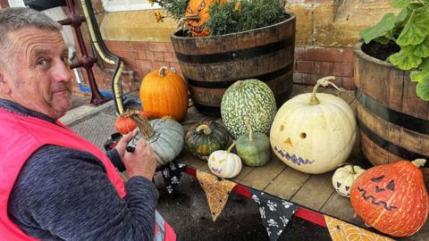 David Rose kneeling down by a railway trolley with autumn vegetation on it, including pumpkins. He is wearing a pink high-vis jacket. There are two wooden planters, with plants in them, on his trolley. 