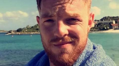 Josh Clayton is seen smiling at the camera with a beach and sea behind him. 