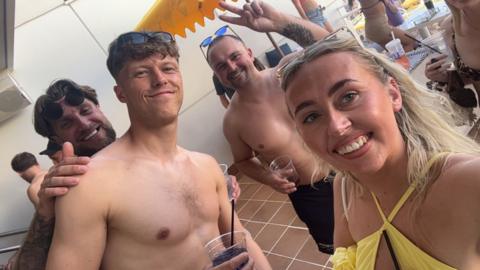 Three bare-chested men wearing swim shorts smiling at a camera held by a woman with blonde hair wearing a yellow bikini smiling