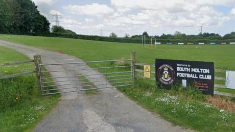 Football pitch at South Molton Football Club. Green pitch. Grey gate and black football club sign. Grey drive going into the site