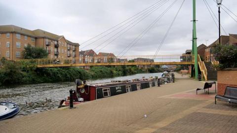 Canal path near Trenery Way in Northampton. There are canal boats in the water by the side of the path and a bridge with yellow railings across the water.