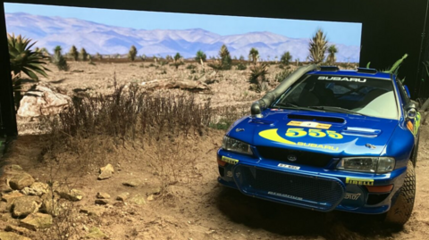A blue rally car with 555 printed written in yellow across the bonnet, positioned indoors on a set of a sand and rocks and a bank of vegetation. At the back is a wide screen showing a similar scene of sand, dry vegetation, palms, and mountains rising in the background. 