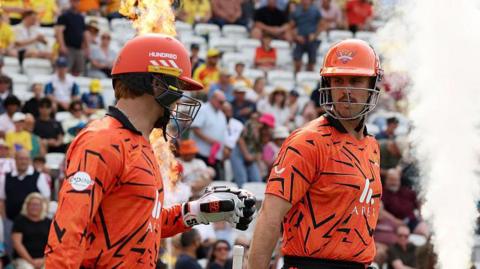 Ryan Rickelton (left) and Mitch Marsh (right), wearing orange shirts and helmets, walk through pyrotechnics onto the field