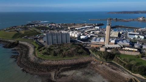 The image shows an aerial drone shot of St Helier. There is a rocky shoreline with some greenery and trees. On land there are several large buildings and an industrial facility with a tall tower. The ocean is visible on both sides with clear blue skies above.