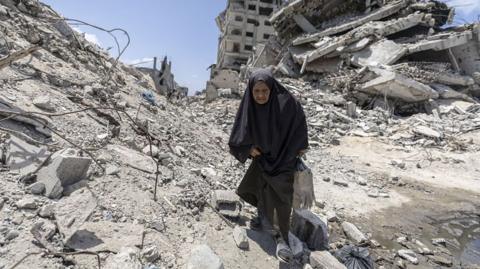 A Palestinian woman steps over rubble in Gazy City, with badly damaged buildings and debris behind her