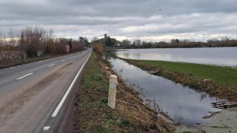 The road, which has some mud on it, is clear of floodwater. The field next to it is almost entirely waterlogged, with only a thin strip of grass visible. Glastonbury Tor can be seen in the distance. 