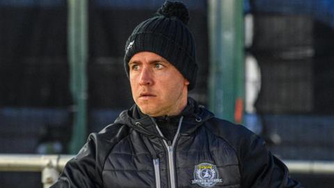 Ritchie Hawkins wearing a black, woolly hat and a black padded zip-up jacket with Ipswich Witches insignia on the chest.