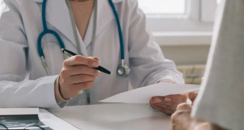 A doctor in a white coat with a blue stethoscope around her neck sits at a desk, photographed from the neck down. They are holding a pen in one hand and a piece of paper in the other.