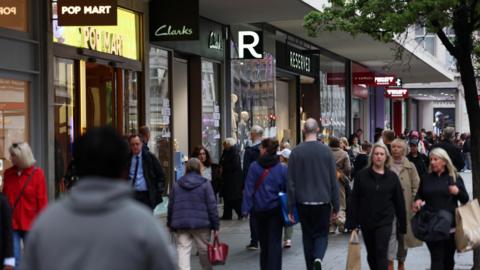 People walk along high-street, some carrying carrier bags. Behind the crowd are signs for the shops Pop Mart, Clarks, Reserved and H Samuel