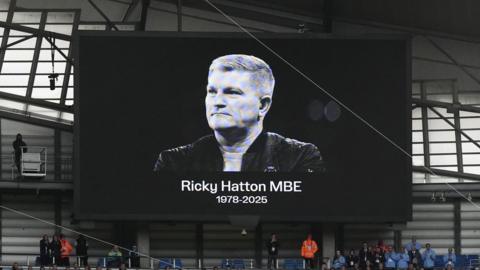 A televised billboard inside a football stadium showing a black and white photo of Ricky Hatton, wearing a black jacket and smiling, above his name and the years of his life, 1978-2025. 