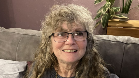 Debbie Crowson sitting on a grey sofa and smiling. She has curly grey hair and is wearing glasses with black rims.