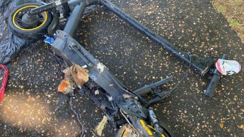 A black e-scooter laid on its side on a road. It has partially crumpled up due to fire damage and has rubbish stuck to parts.