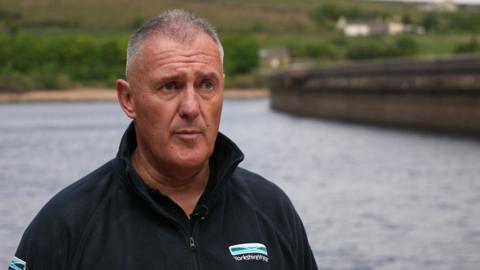 A head-and-shoulders image of Dave Kaye. He is a middle-aged man with short grey hair and is wearing a black zip-up fleece featuring the Yorkshire Water logo. Mr Kaye is standing in front of a reservoir.