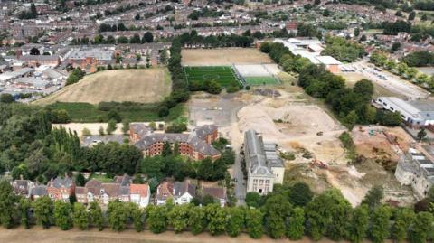 The campus land in a aerial shot shows a number of buildings and land that has been dug up and there are some fields and a sports pitch in the background.