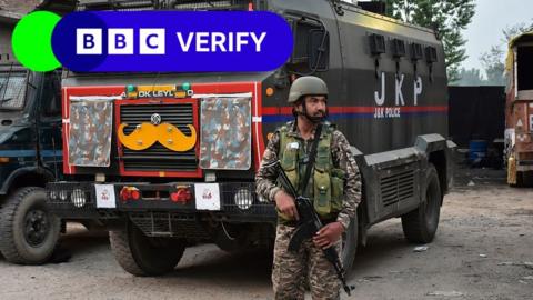 An Indian soldier stands guard. He is armed with a rifle and is wearing army fatigues. In the background is a large truck.