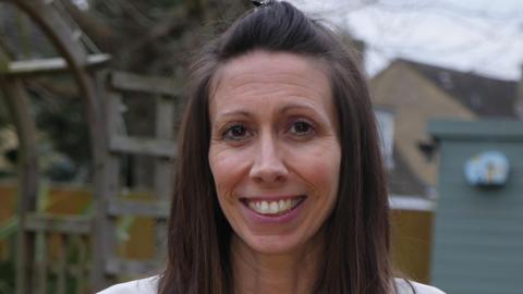 Kristie Higgs smiles at the camera. She has long brown hair down to her shoulders and is wearing a white top