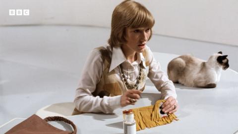 Lesley Judd sits at a white desk, holding a Dougal made from felt. A cat sits on the side of the desk