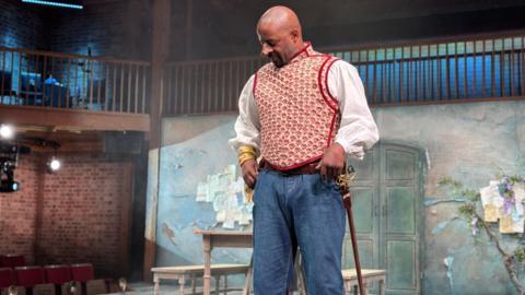 A man standing on a stage during a performance of a play. He has an enhanced, disfigured nose, a red and white waistcoat over a white shirt, a sheathed sword, jeans and is looking down with his hands at his pockets. Behind him are tables and chairs, as well as a balcony.