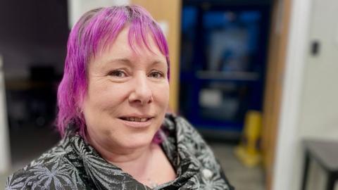 Leanne Nagel poses close to the camera. She has dyed purple hair and is wearing a black patterned blouse.