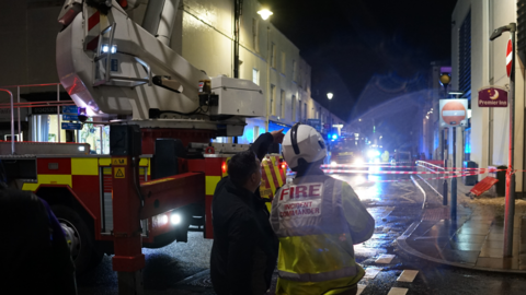 A firefighter is seen from behind as he looks onto a closed road with two fire engines attending.