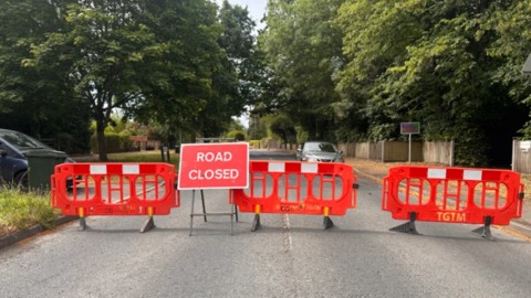 A red road closed sign in front of crash barriers on a residential street, with a silver car parked behind them. A blue car is to the left of the picture.