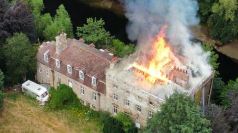 Overhead shot of Armathwaite Castle, a large mansion with three floors. The wing to the left is shorter and includes dormers on the top floor, while the roof of taller wing to the right has flames are shooting out of the middle of its roof. The building is surrounded by trees and a lawn and there is a large caravan parked outside.