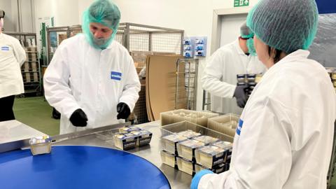 Together a man and a woman, wearing green hair nets and white overalls, are picking up tubs of clotted cream off the conveyor belt to be placed in trays at the end of the line