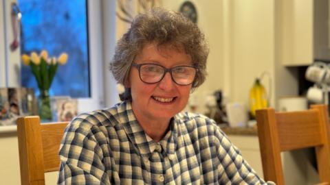 A woman with curly grey hair and black glasses is sitting in a kitchen and smiling at the camera. She is wearing a blue and white checked shirt.