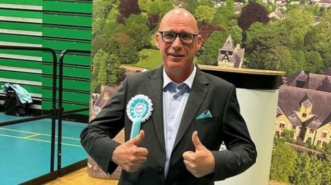 Anthony Owen in a jacket and shirt with a Reform UK rosette, giving a double thumbs-up
