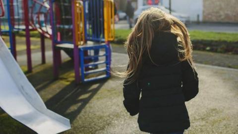 A general view of a play area with a slide and climbing equipment in bright colours. A young girl has her back to us, wearing a dark coat and with long, brown hair past her shoulders