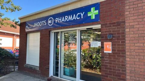 In bright sunlight, the photo shows the front of the Jhoots branch in Victoria Square, Portishead. Above the door a giant blue banner featuring NHS branding reads "Jhoots Pharmacy". To the left of the door a window has shutters pulled down in front of it. 