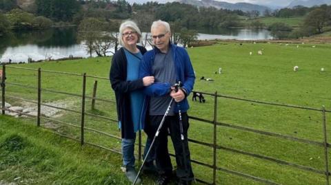 A woman and a man are standing alongside a metal fence with a field and large body of water on the other side. There are sheep in the field and in the distance is woodlands and further in the distance is a range of hills.