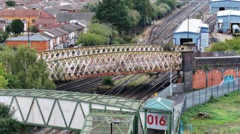 A high view looking down on a rail bridge above the tracks. Its is white and red. Houses can be seen one side of the rail line and engineering units on the other.
