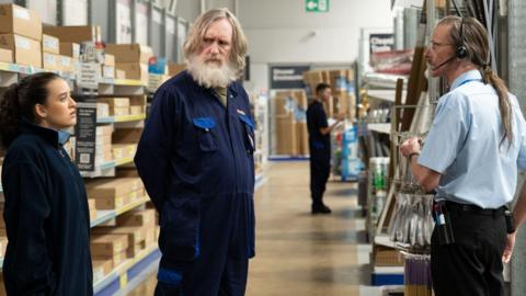 A young woman and an older man with a beard and long hair stand wearing jumpsuits in a DIY shop. They are speaking to a thin man with long hair and wearing a button-up shirt tucked into trousers and a headset. 
