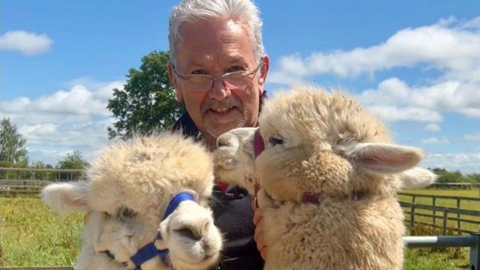 A man with white hair and glasses is posing with two fluffy cream coloured alpacas. Behind them is a wooden fence, blue sky, large tree and a field of green grass.