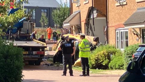 A car on its side in front of a house which it has crashed into. Part of the front wall of the house has collapsed. There are two police officers standing by the scene with a recovery vehicle.