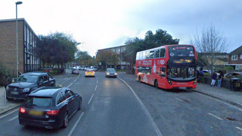 A double decker bus is stopped at a bus stop near Templars Bar on Barns Road. A police car and two private vehicles are passing.