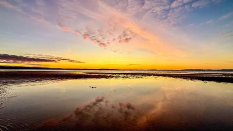 Yellow horizontal lines and pink clouds fill the sky as the sun sets across a bay